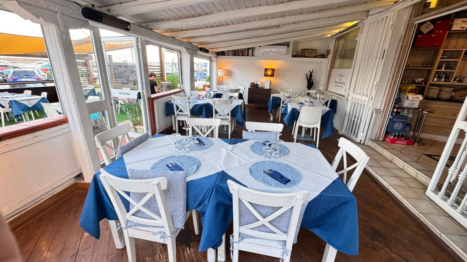Terrazza esterna del ristorante con tavoli e sedie blu e bianchi e pavimento in legno sotto una tettoia bianca.