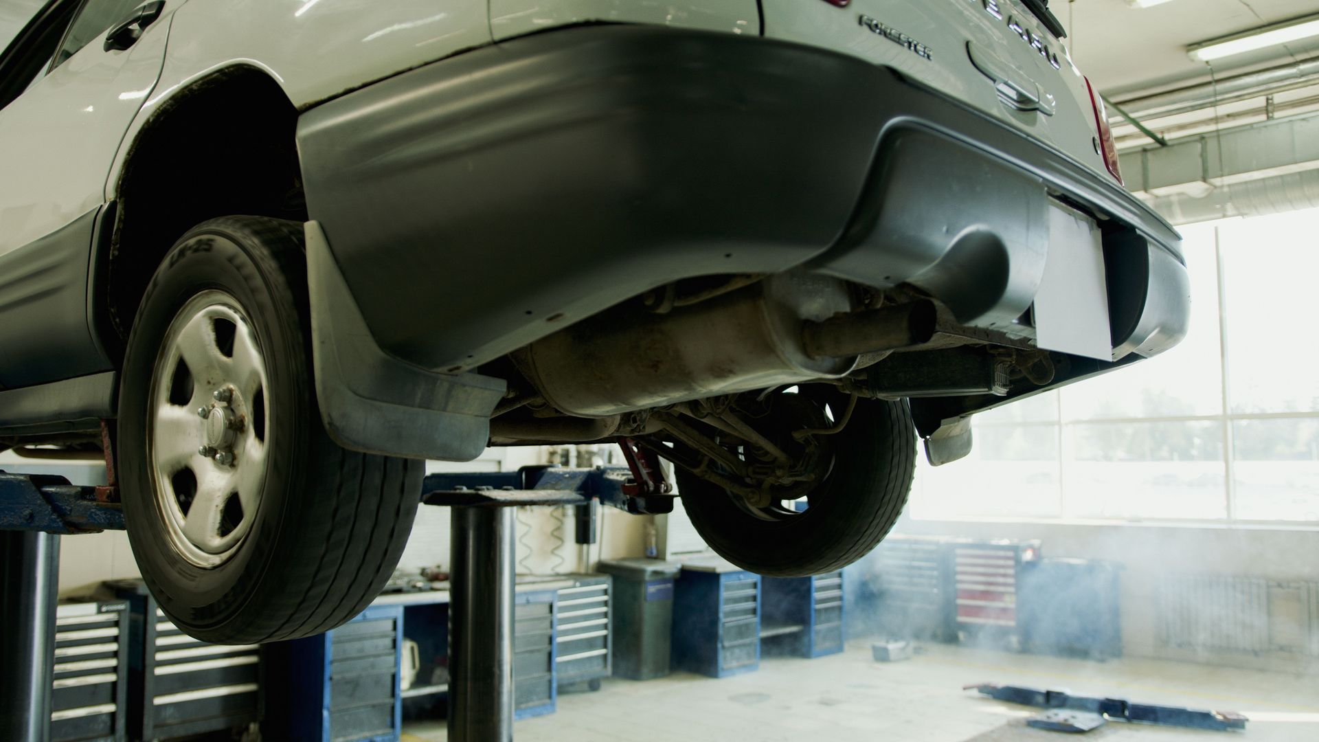 The underside of a car is sitting on a lift in a garage.