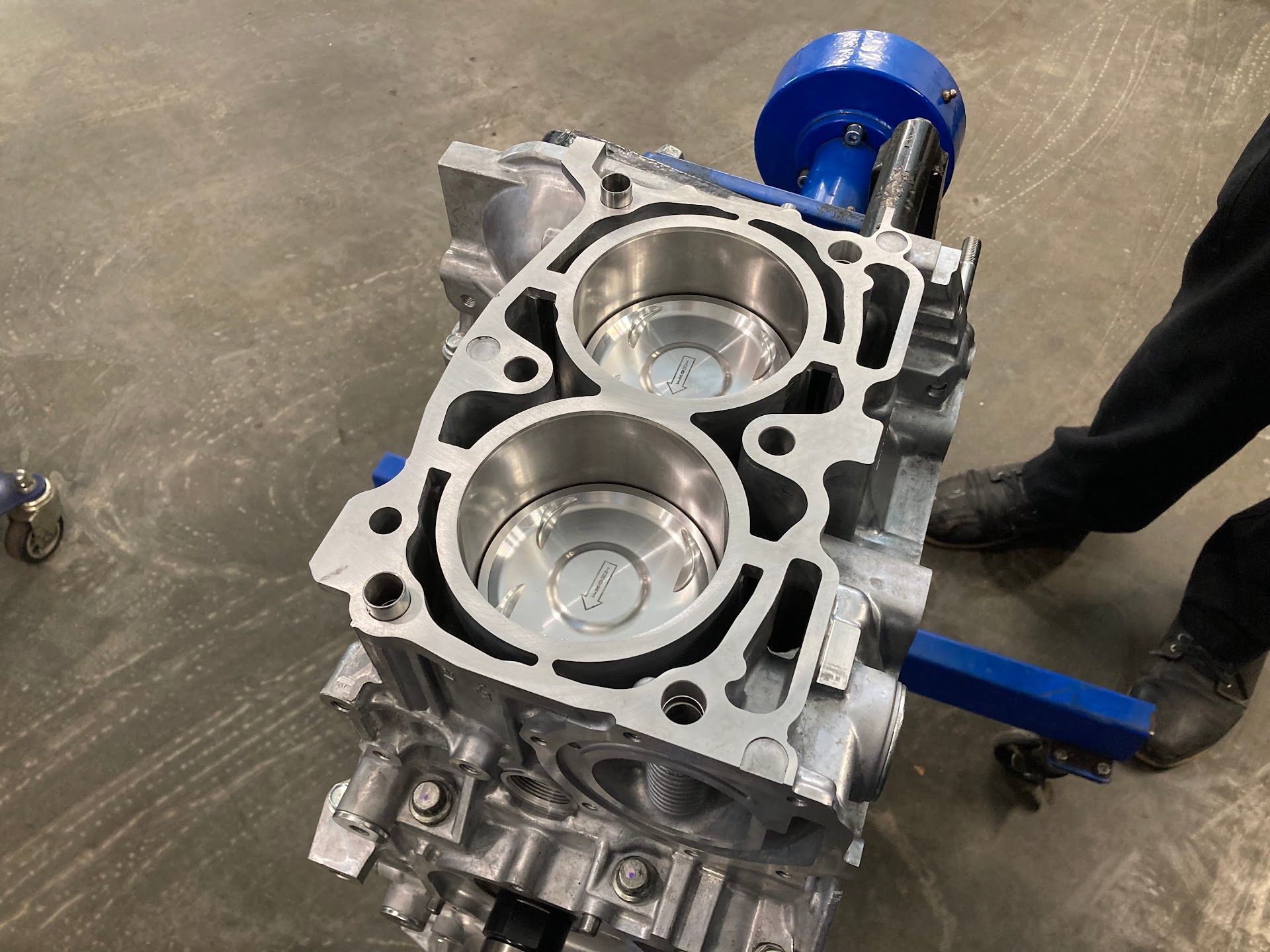 An aluminum engine block mounted on a blue stand in a workshop, showing two exposed cylinder bores. | Teter Automotive