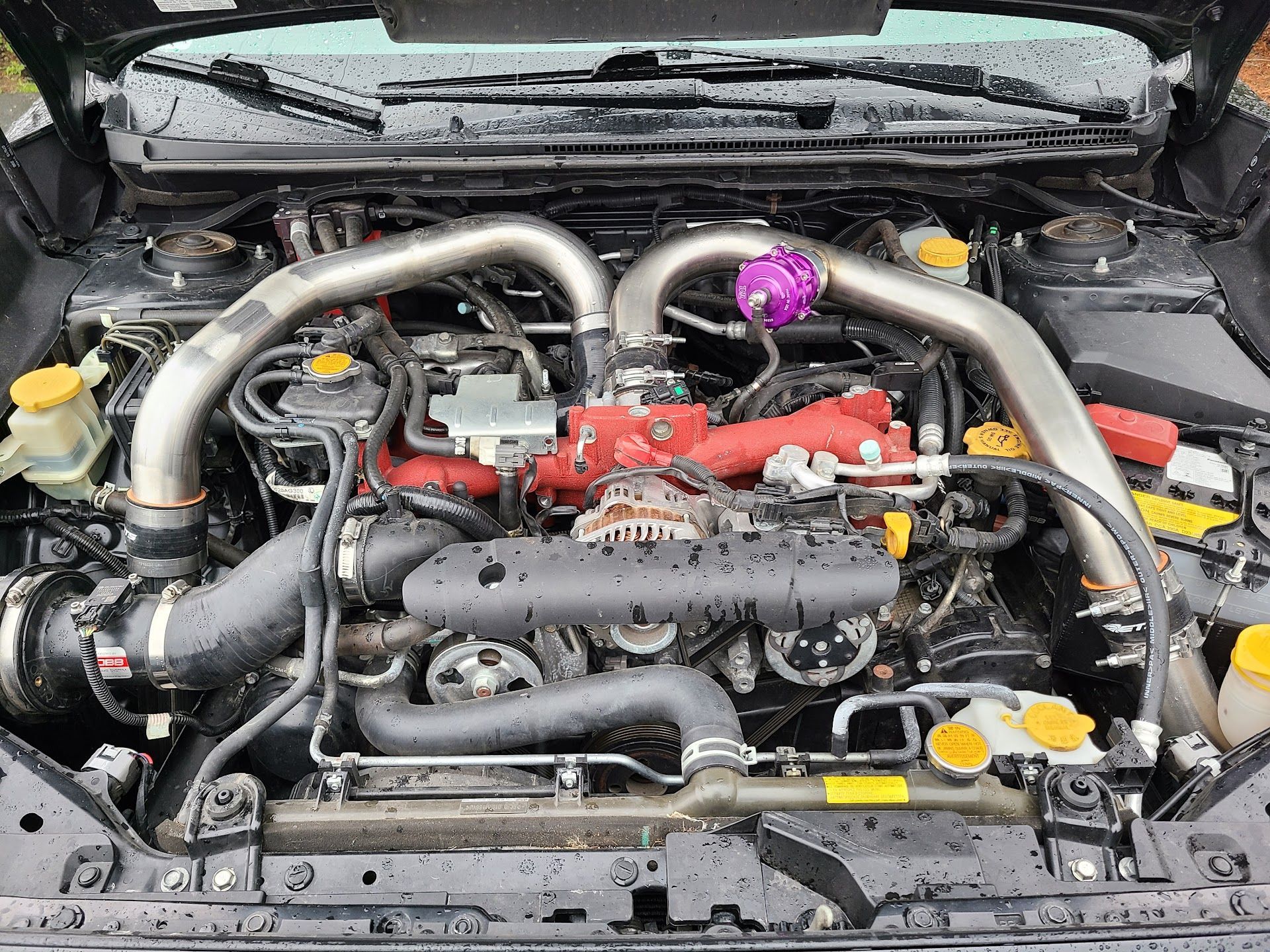 An open car hood showing a Subaru engine bay with a prominent red intake manifold, metallic piping, and a purple part. | Teter Automotive