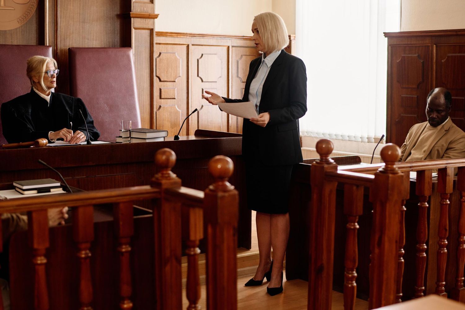 Lawyer speaking in a courtroom beside a judge, with two seated attendees in the background