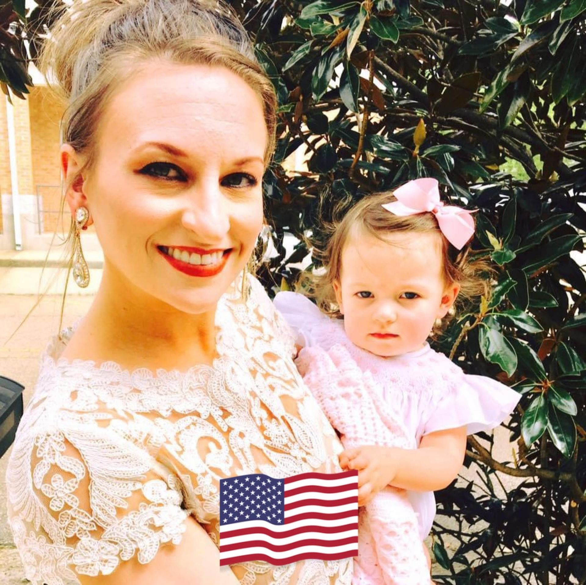 Woman holding a child in pink, both smiling outdoors with a small American flag in front