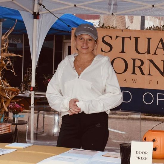 Woman in white shirt and blue cap at a booth with a Stuart Corn sign, standing behind a table.