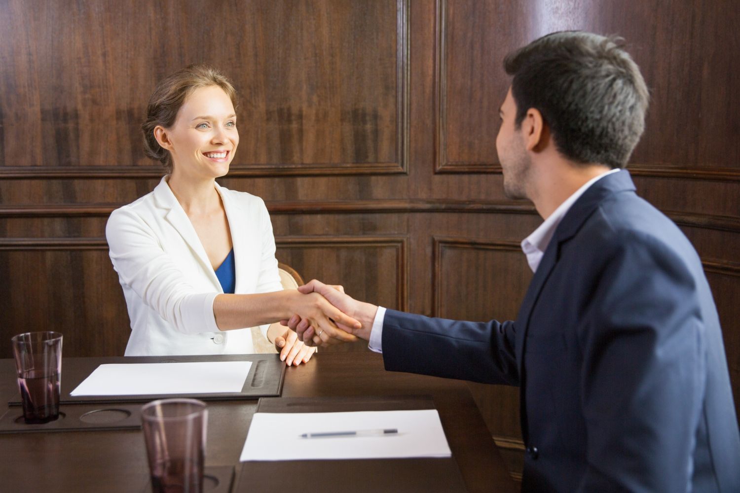 Two people shaking hands across a conference table in a wood-paneled office