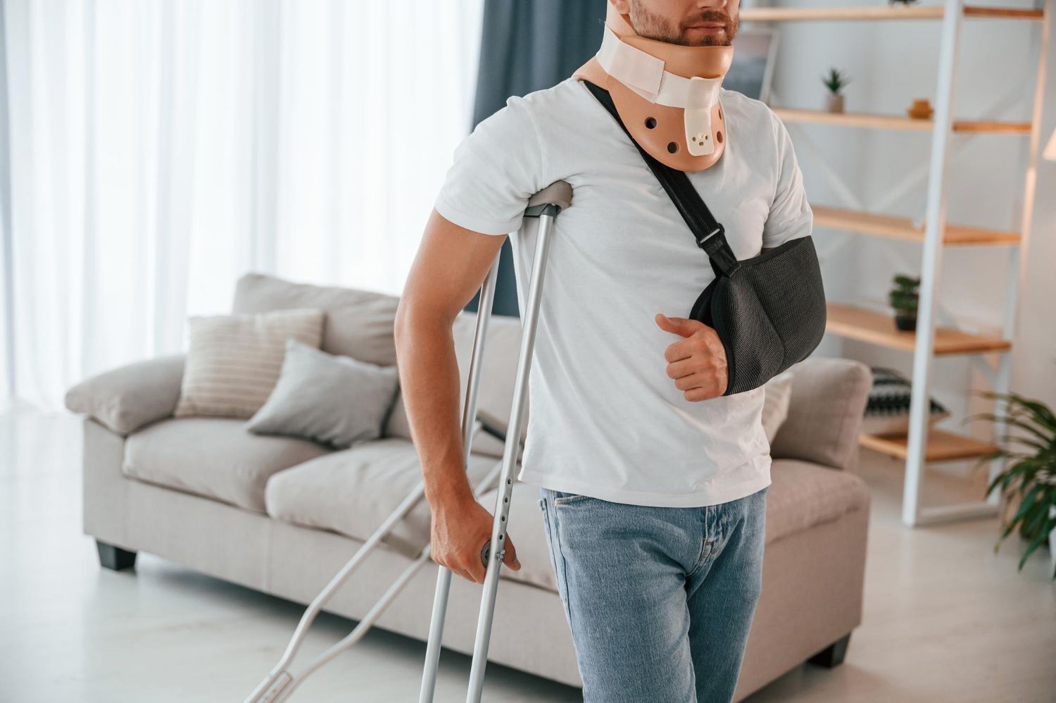 Man with arm in sling and neck brace walking with crutches in a bright living room