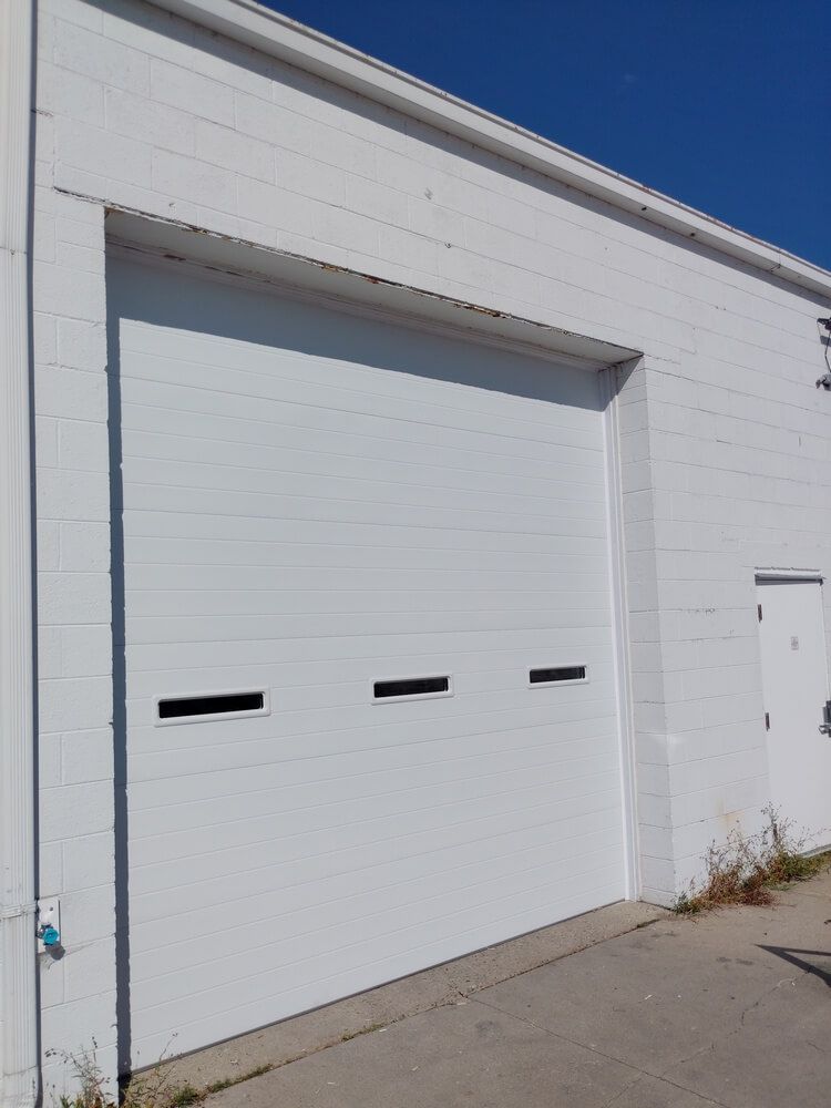 White industrial garage door in a sunlit brick building with a small side door and clear blue sky.