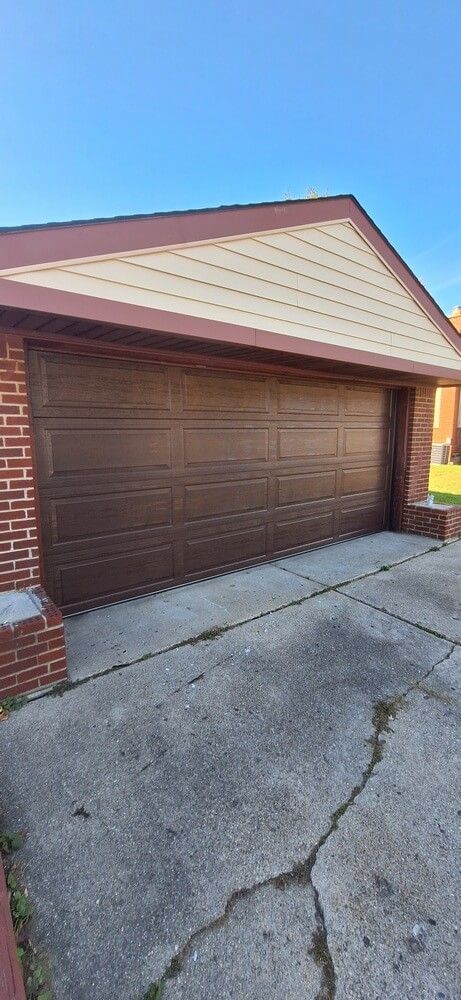Residential garage with brown door, beige siding, red brick trim, and cracked concrete driveway