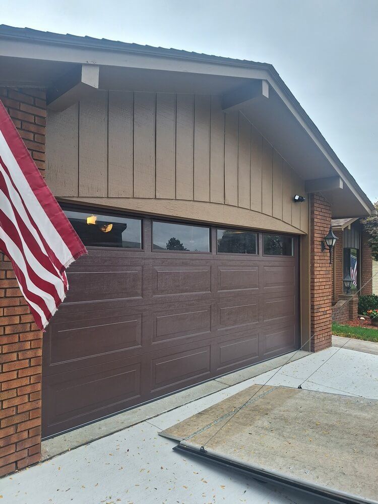 Brown garage door with beige siding, brick wall, and an American flag by the driveway