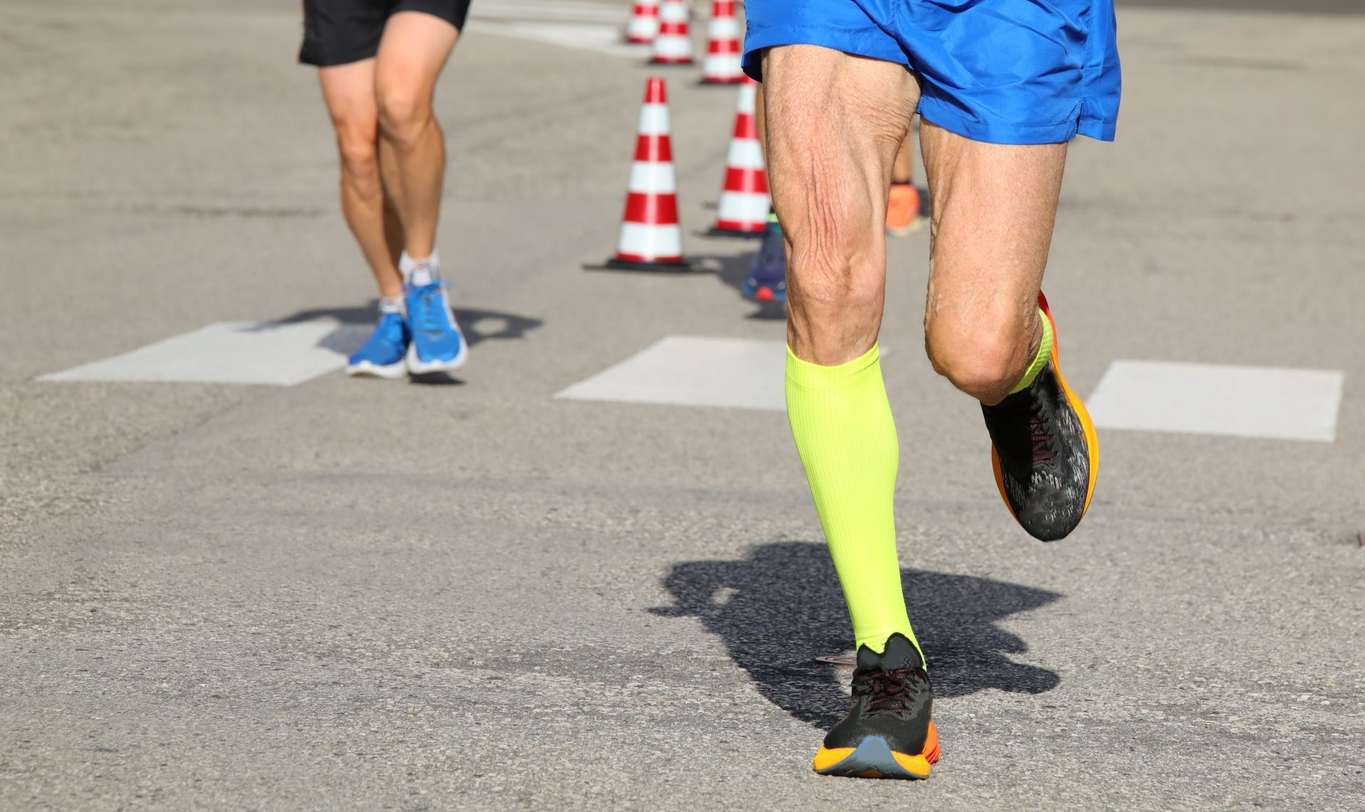 Two runners' legs in action on a paved road, orange traffic cones, one runner in blue shorts and a neon sock.