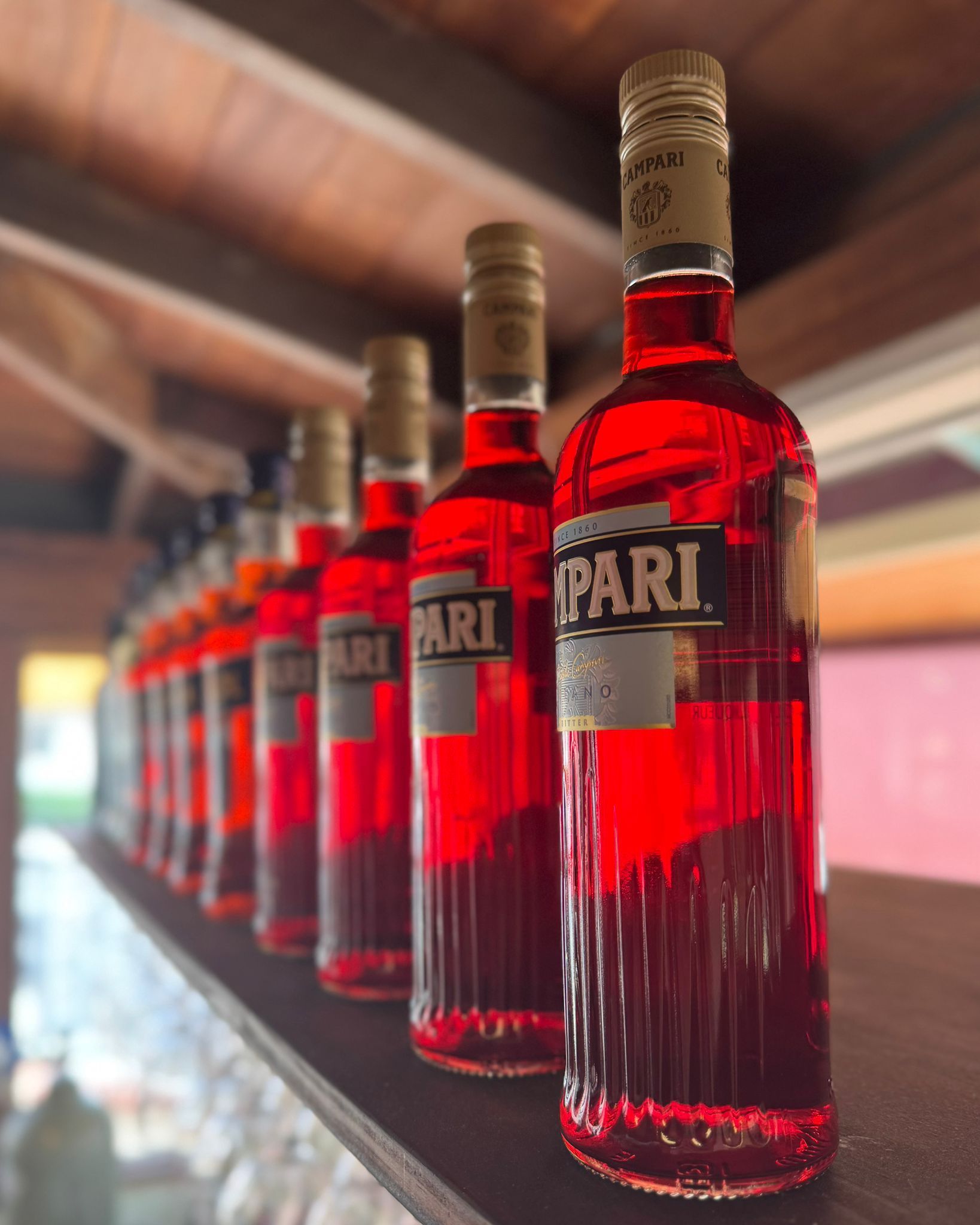Row of Campari bottles on a shelf, vibrant red liquid, blurred background, warm lighting.
