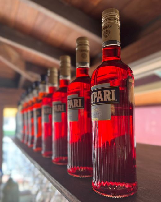 Row of Campari bottles on a shelf, vibrant red liquid, blurred background, warm lighting.