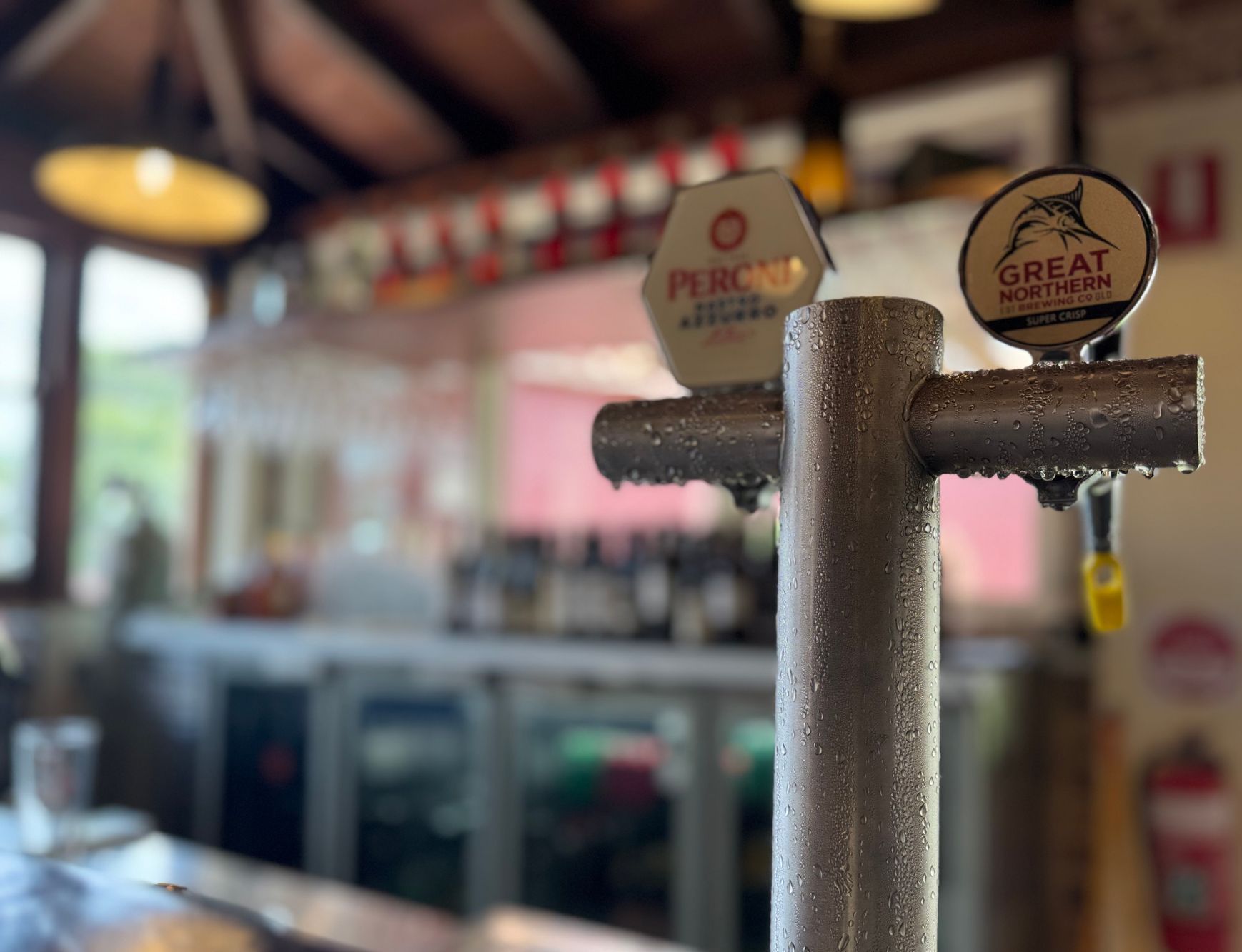 Close-up of a bar tap with condensation, in front of a blurry bar setting with beer taps and glasses.