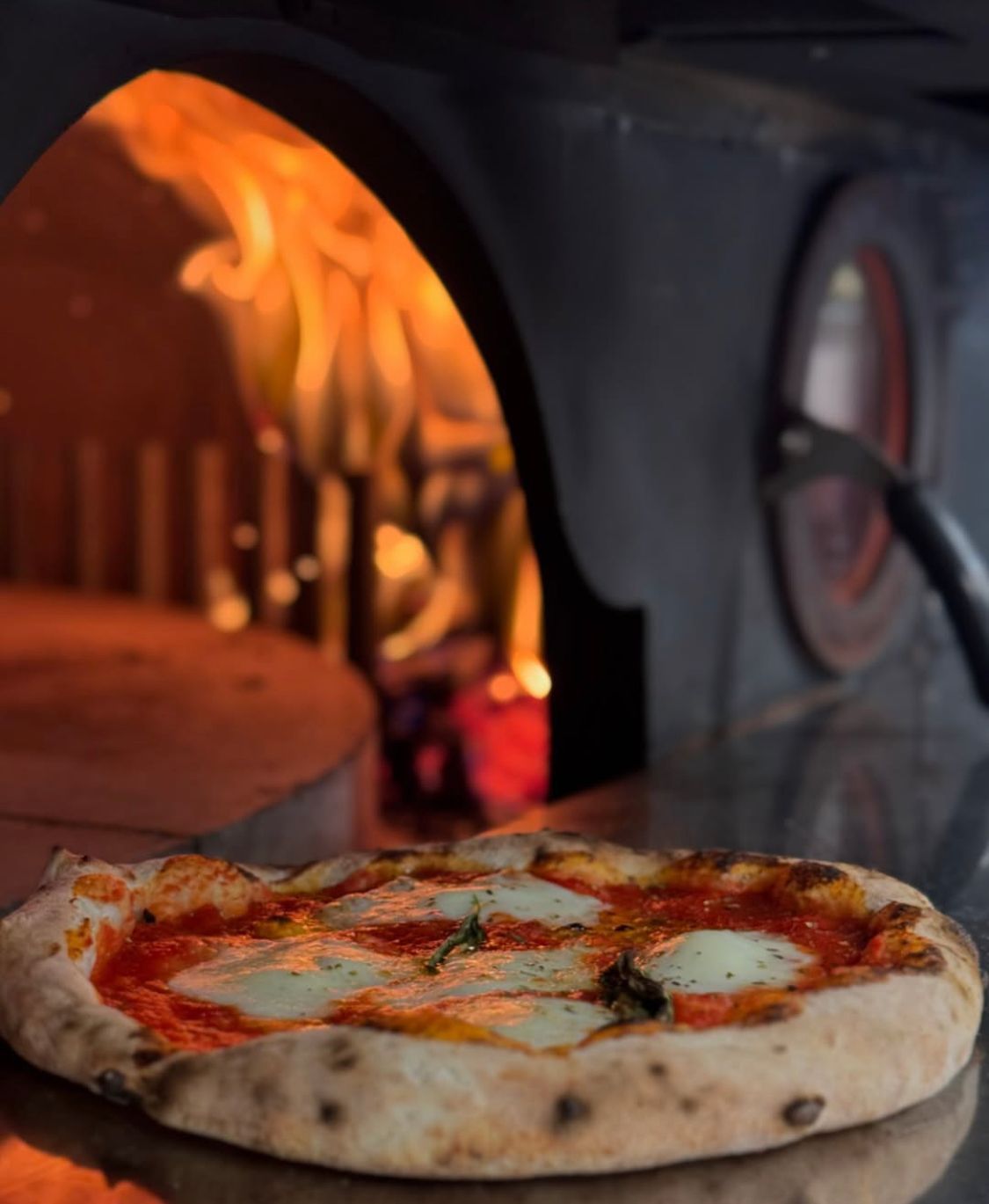 Pizza with mozzarella in front of a wood-fired oven, flames visible.