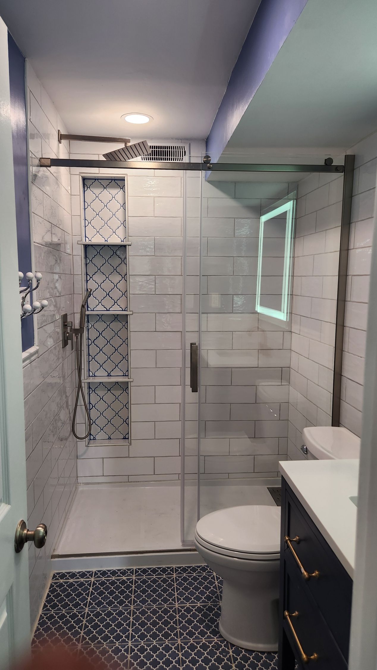 Modern bathroom with glass shower, white tile, patterned blue floor, and vanity mirror.