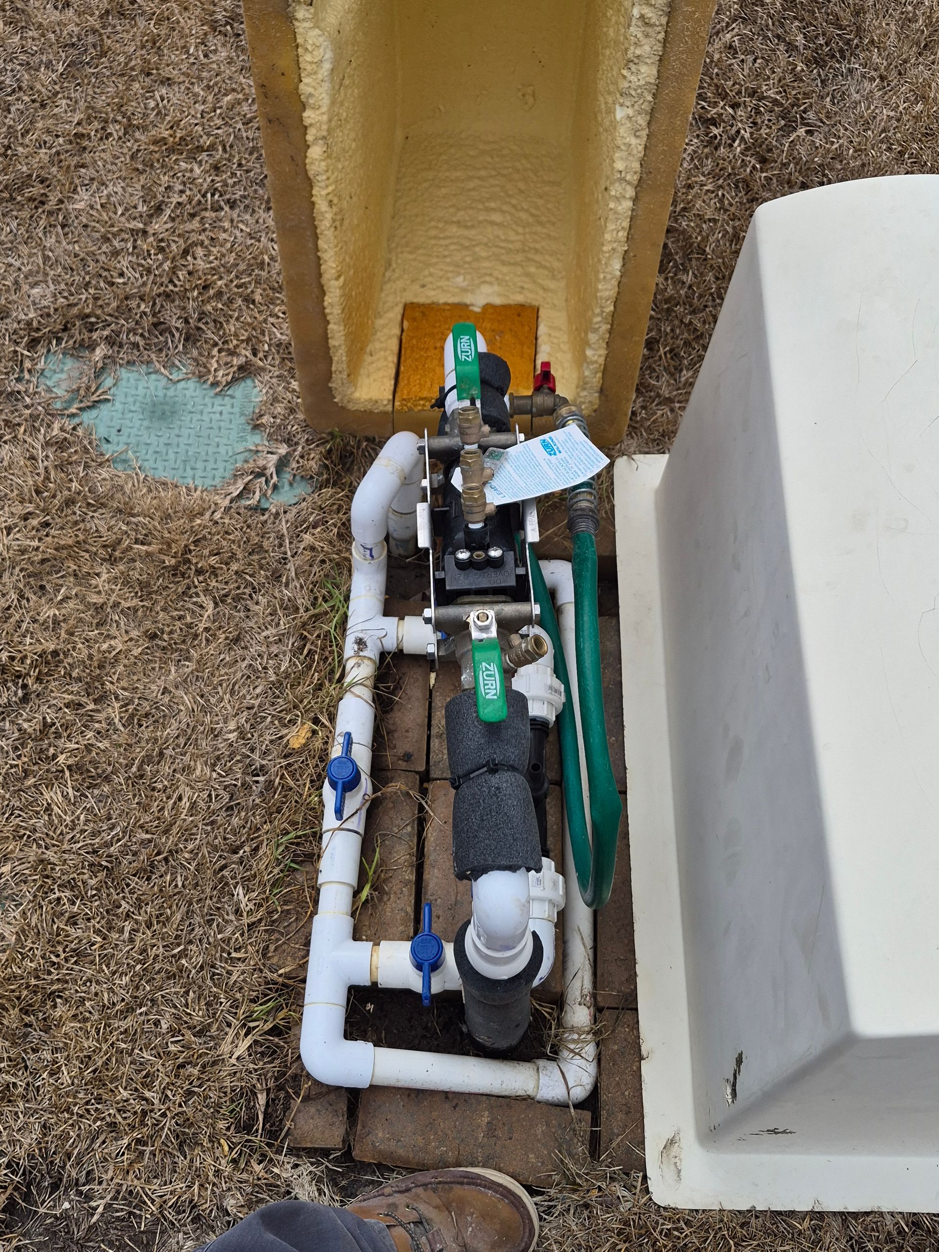 Overhead view of outdoor pipes and valves beside a white building and yellow wall, with dirt and gravel ground