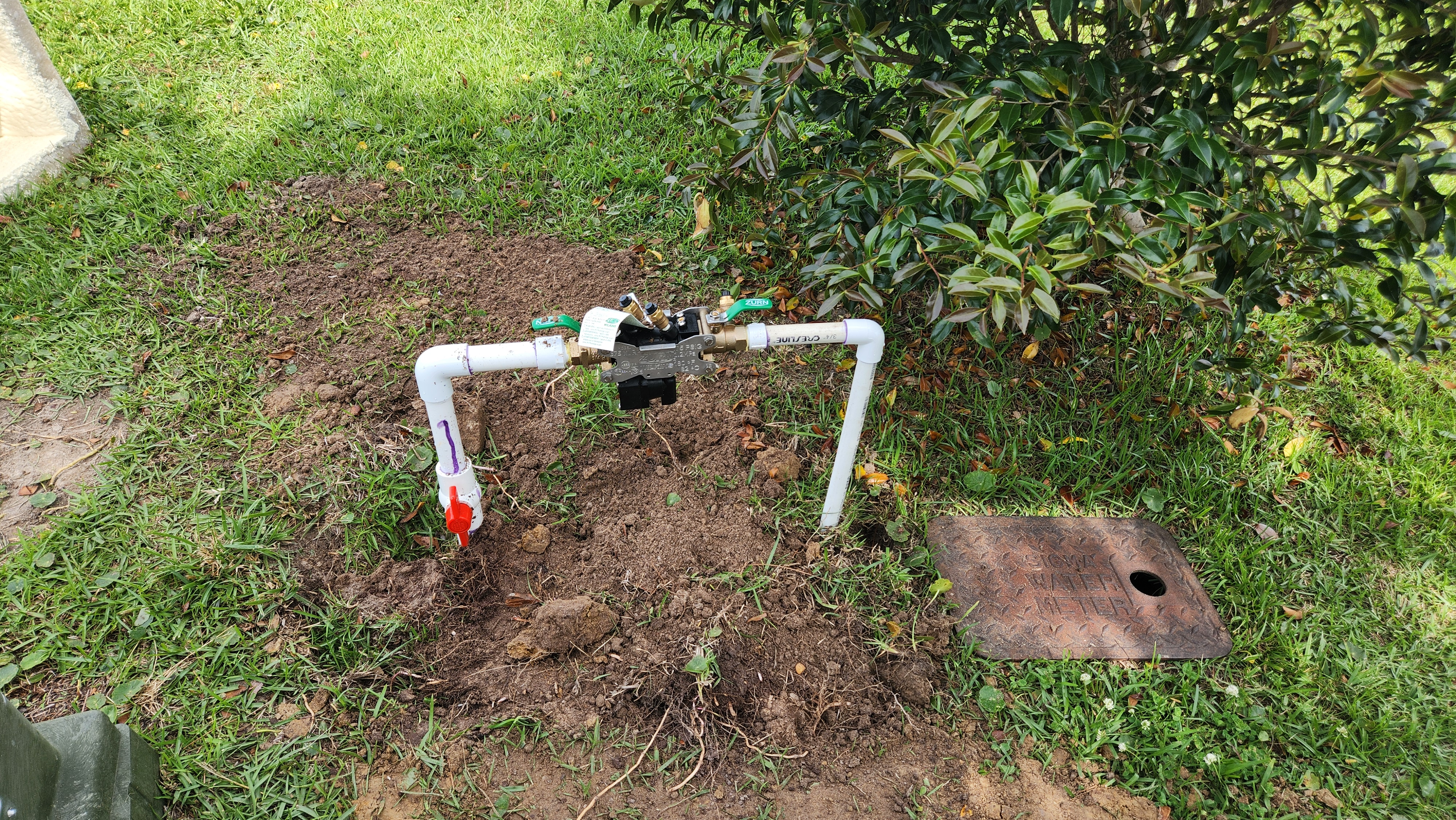 White PVC pipes and a valve on dirt beside greenery, with a red cap and metal plate nearby