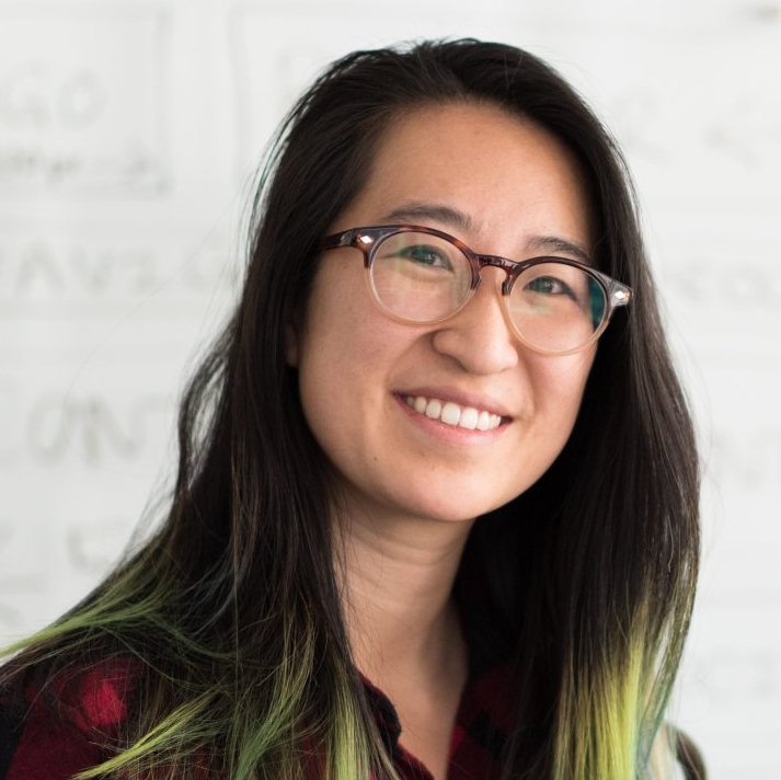 A woman wearing glasses is smiling in front of a white board