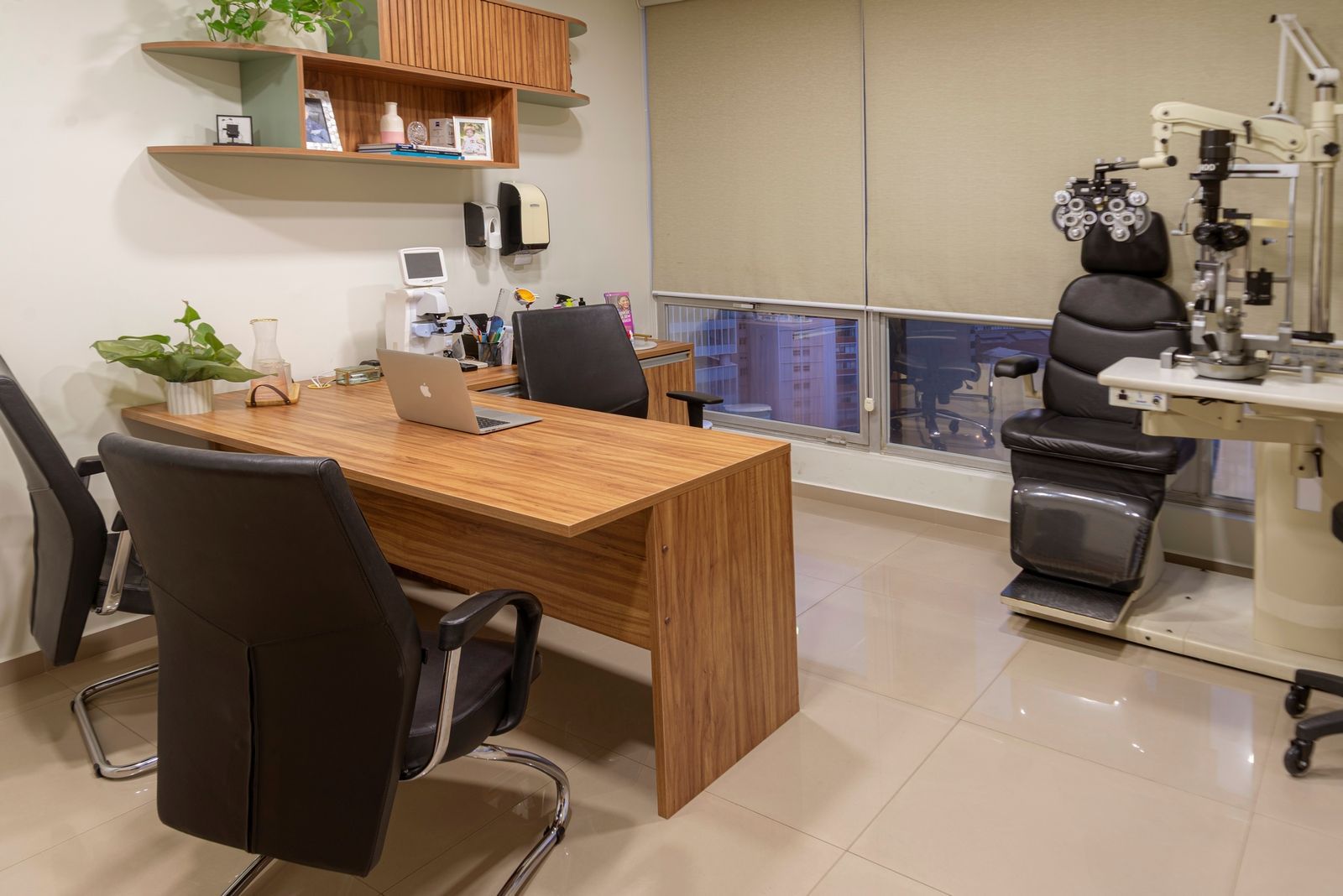 Consultório oftalmológico com cadeira de exame, mesa e equipamentos. Sala bem iluminada com vista para a cidade.