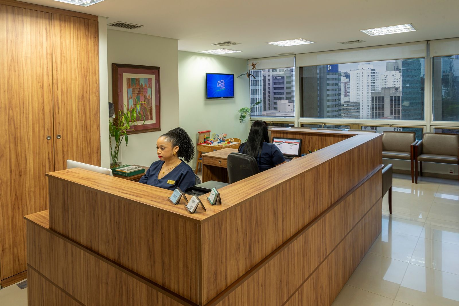 Duas recepcionistas em uma mesa de madeira em um consultório médico, com vista para a paisagem urbana.