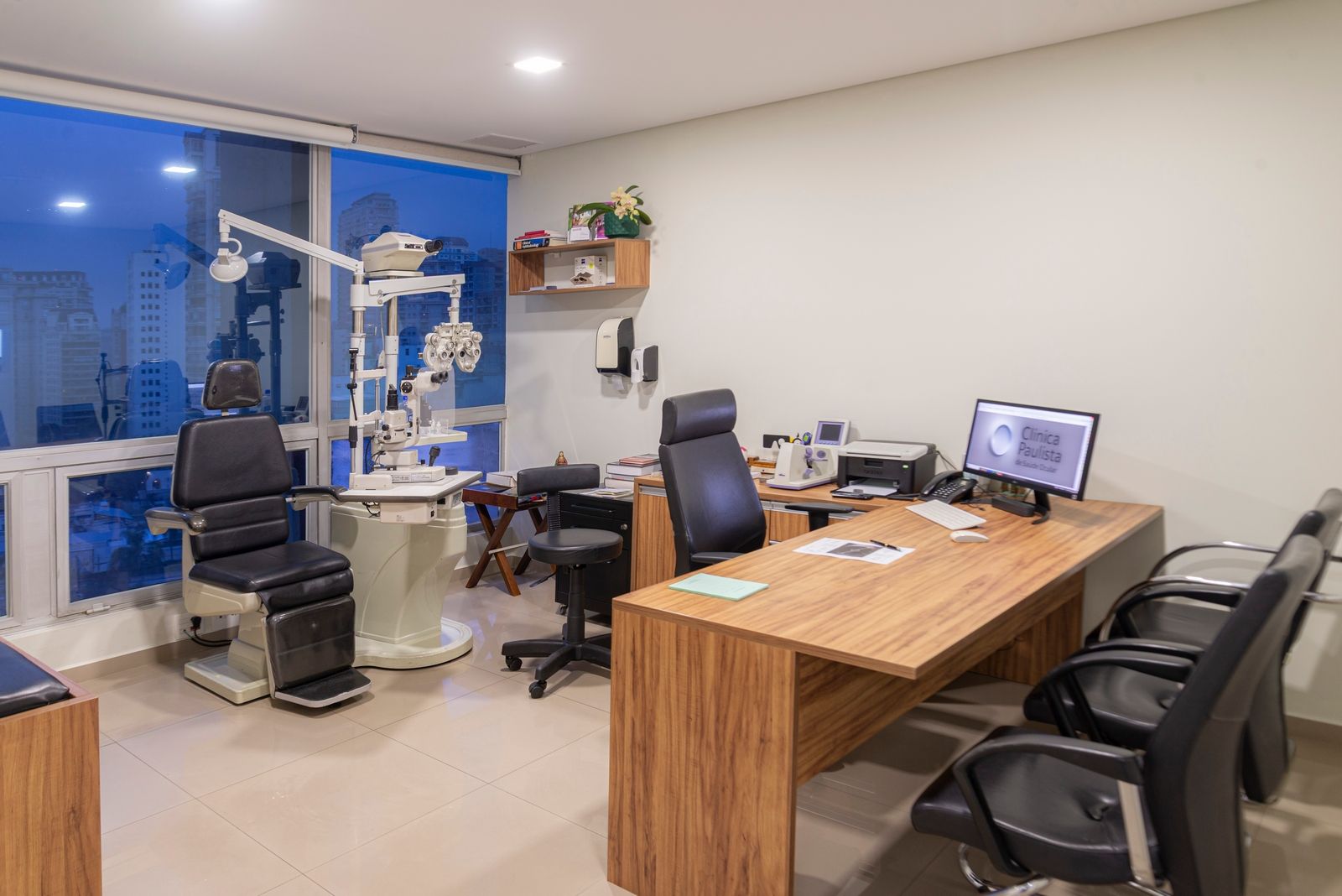 Consultório oftalmológico com equipamentos para exames, mesa e cadeiras. Janela com tonalidade azulada.