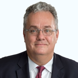 Studio headshot of a man in glasses, dark suit, white shirt, and red tie, against a light background