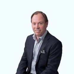 Man in a dark blazer seated against a light background, facing the camera with a neutral expression