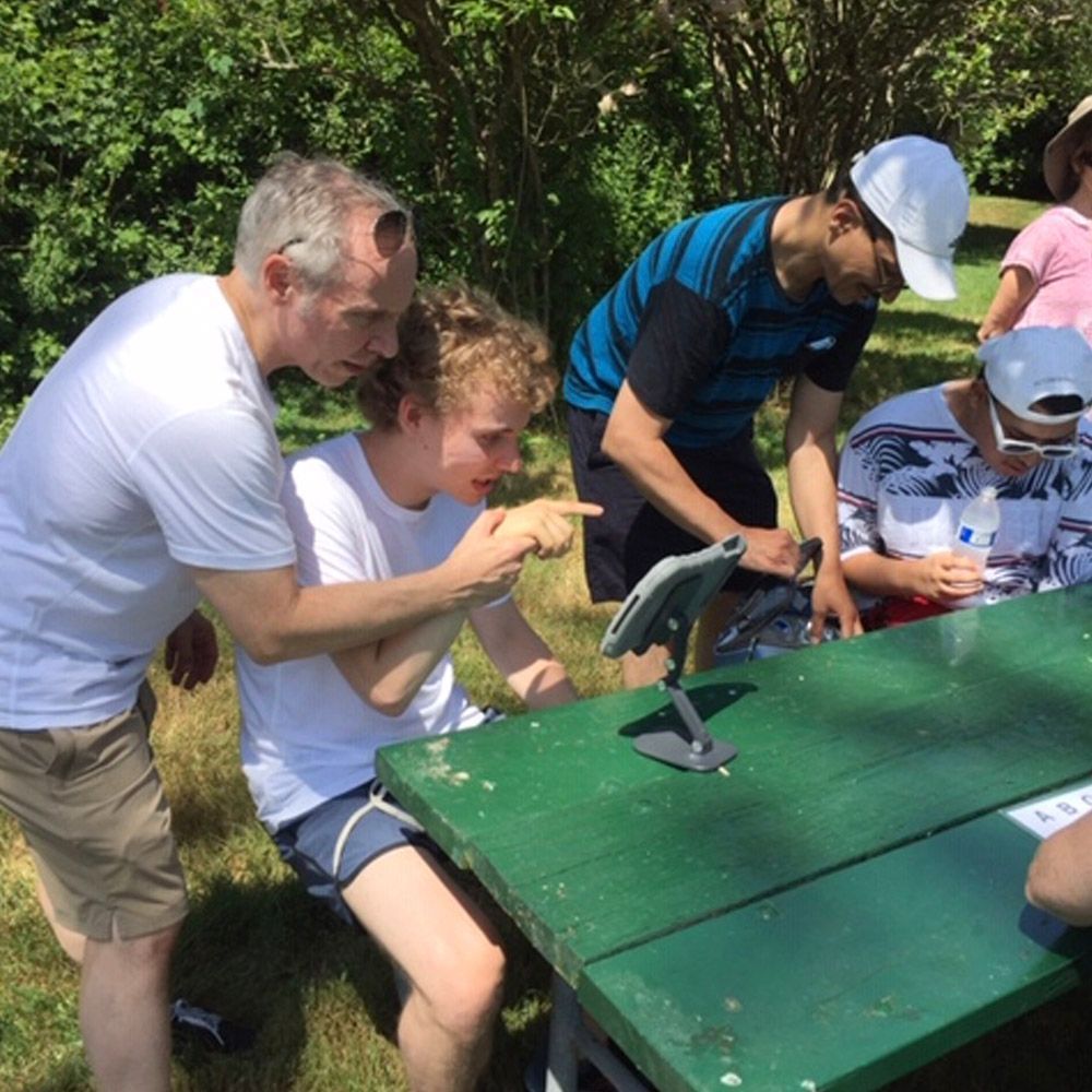 Group of people looking at tablet outdoors, one person pointing. Green picnic table, sunny day.