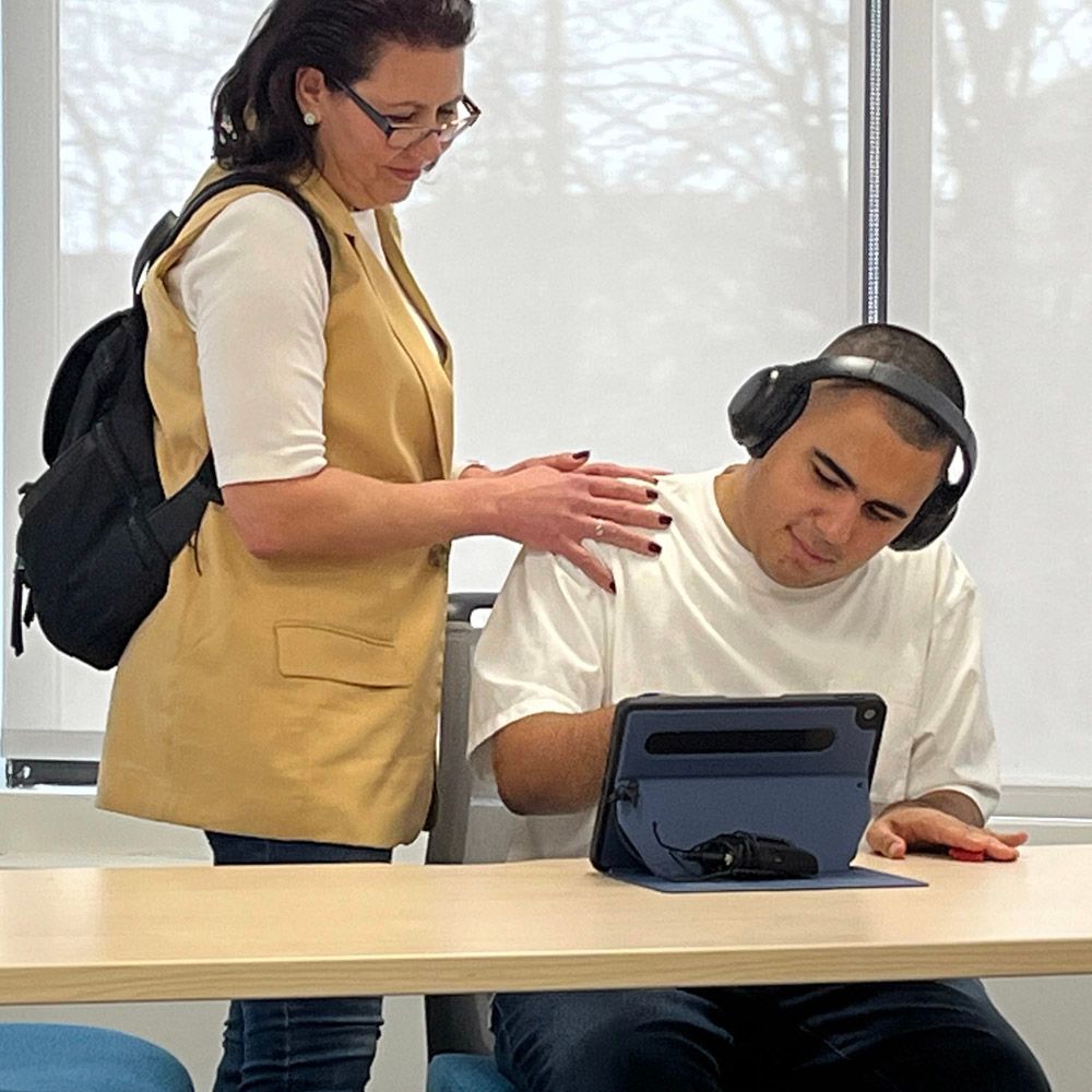 Woman with backpack supports a young man wearing headphones, using a tablet at a desk.