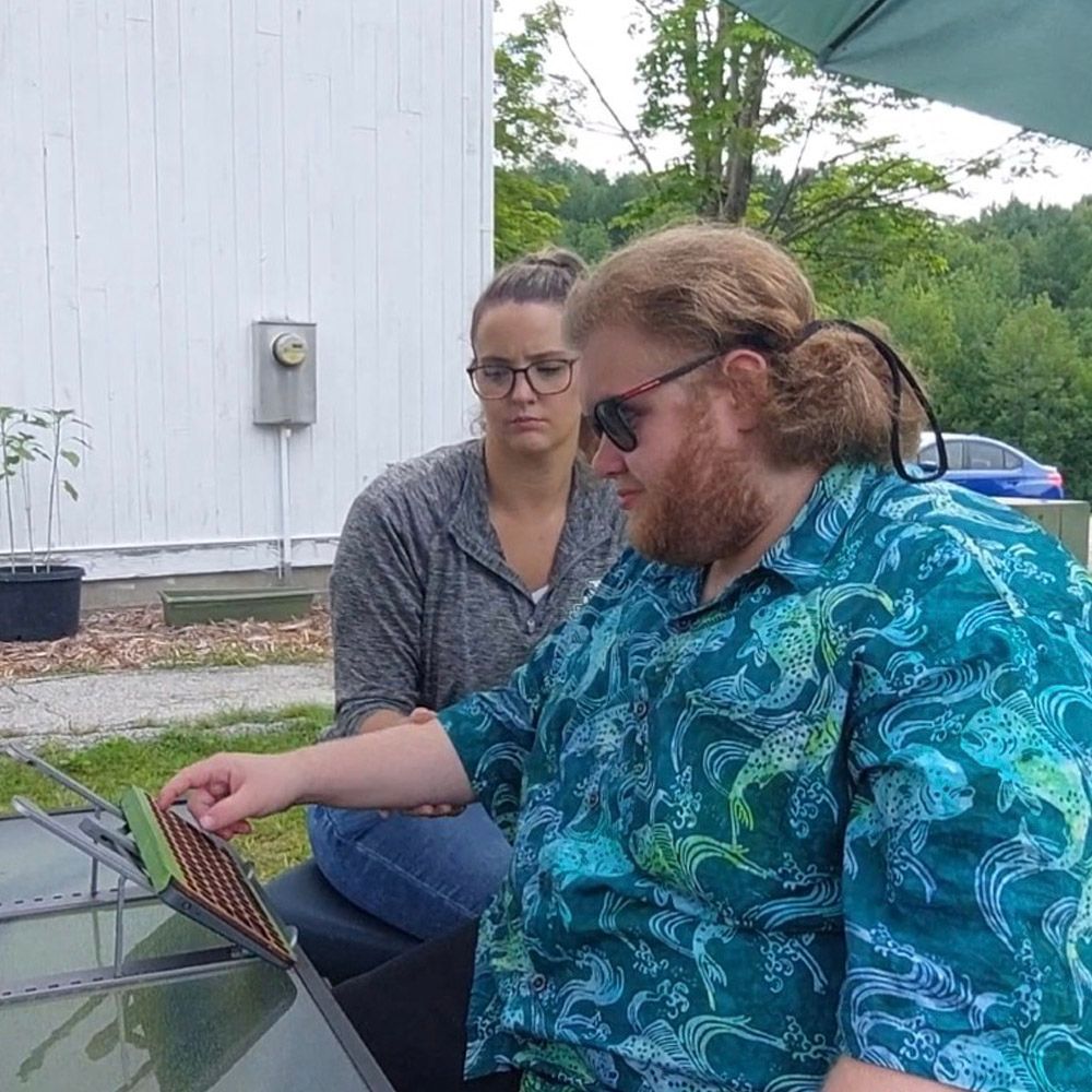 A woman watches a man with a beard inspect a panel outdoors. White building and trees in background.