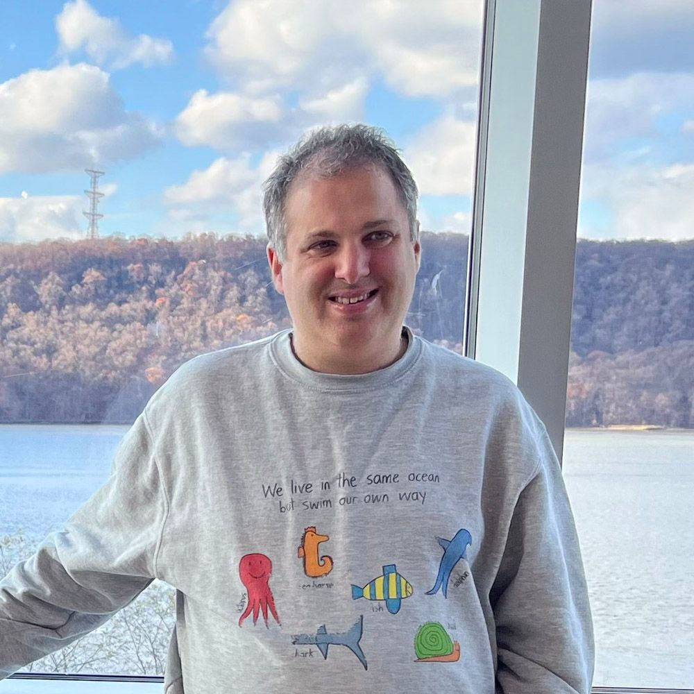 Man smiling in grey sweatshirt with sea creature designs, standing by a window overlooking a river and treeline.