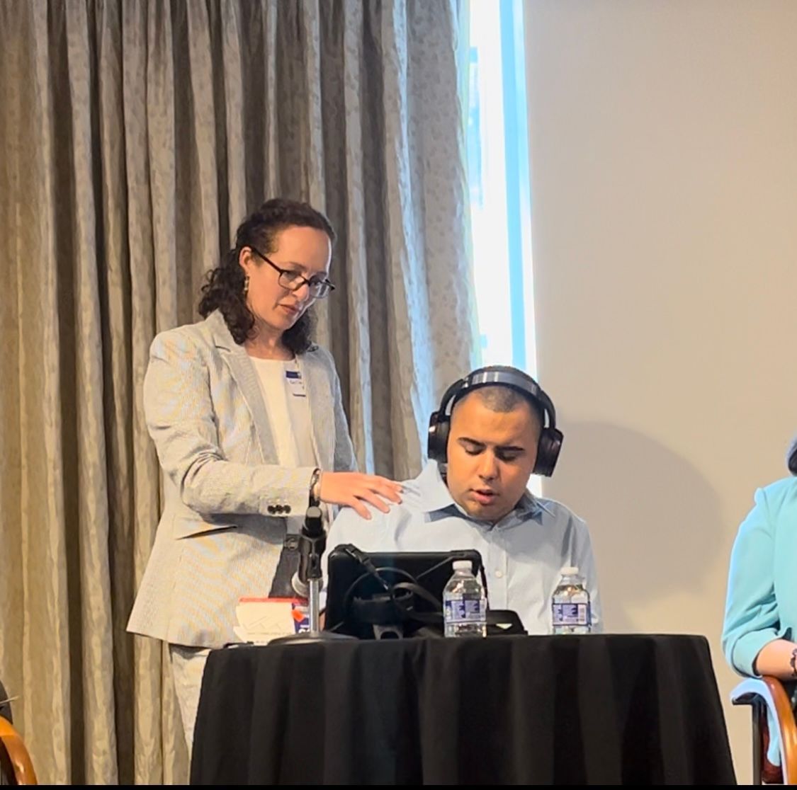 A woman in a blazer assists a man wearing headphones at a table. Conference setting.