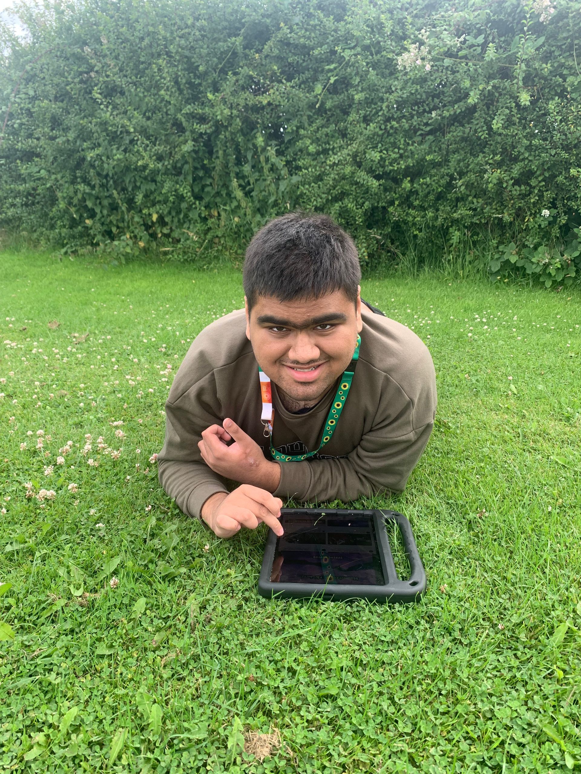 Person with dark hair using a tablet while lying on green grass, smiling.  Green foliage in background.