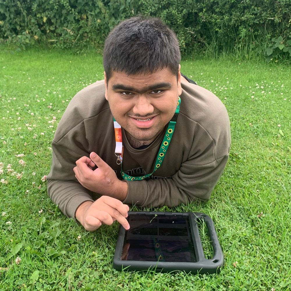 Person with dark hair, smiling, using a tablet on a green lawn. Green lanyard visible.