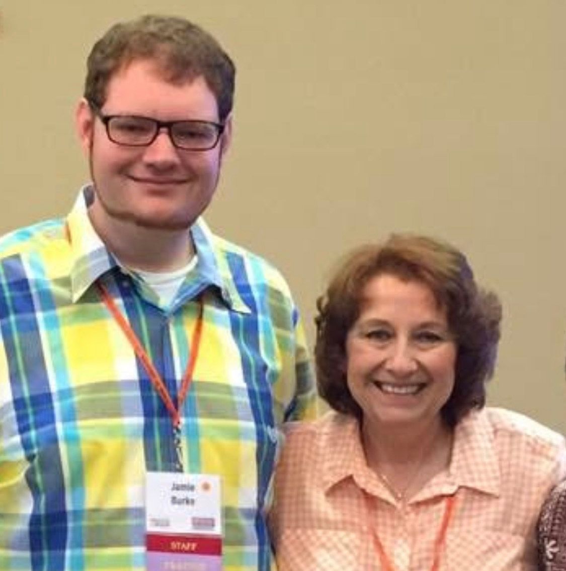Man with glasses and woman smiling; both wearing lanyards, standing indoors.
