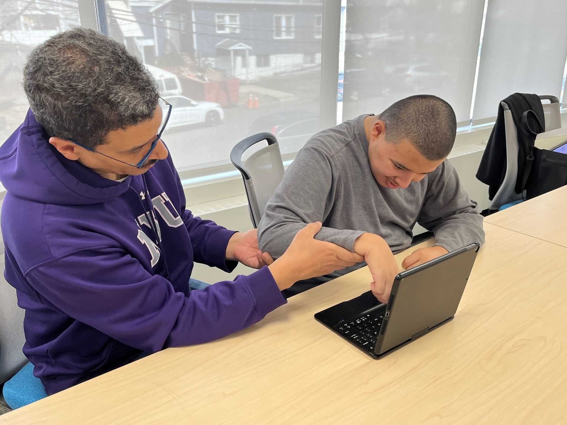 Woman in purple hoodie assisting a man using a laptop at a table, both looking at the screen.