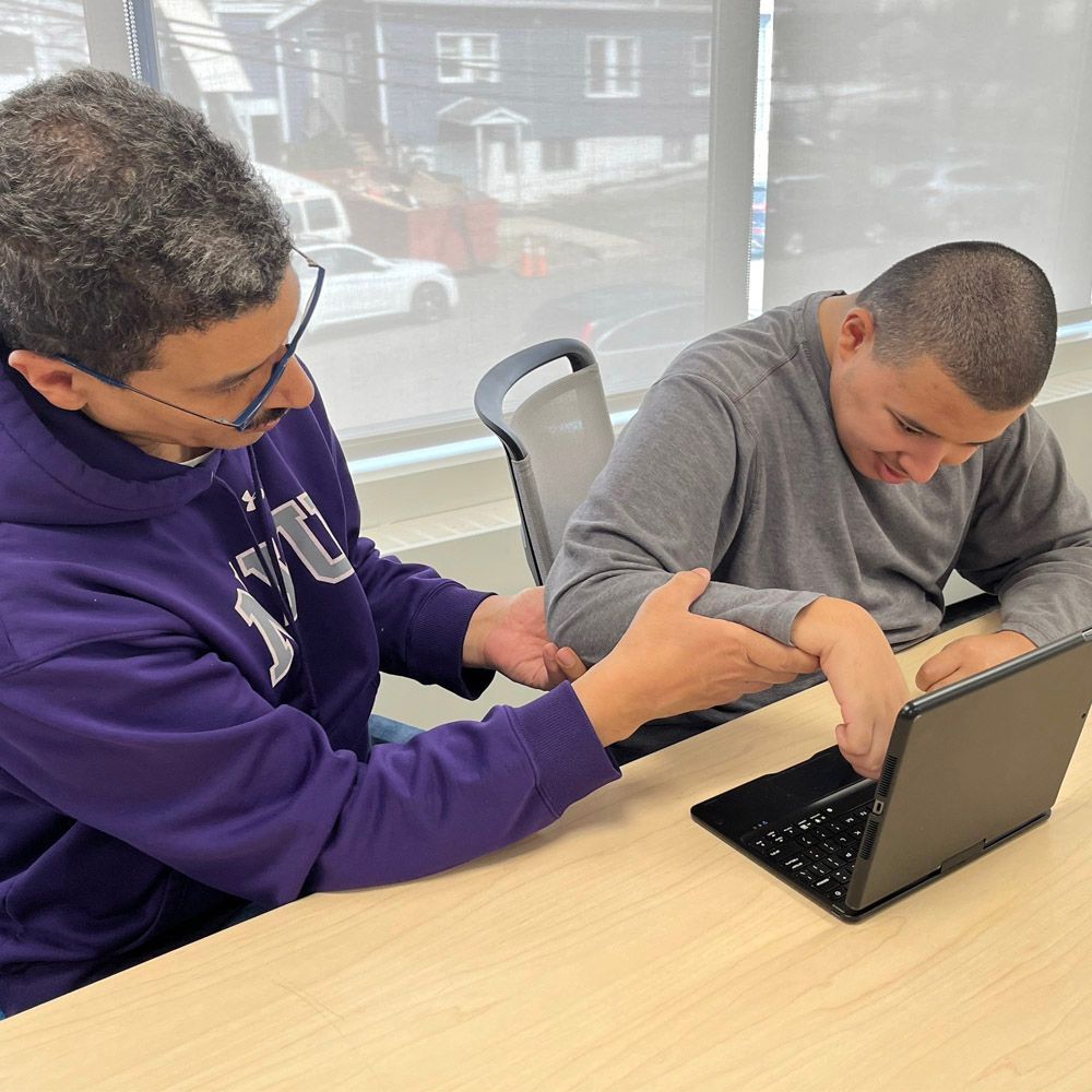 Two people at a desk, one helping the other use a laptop. The helper wears purple. Window in background.