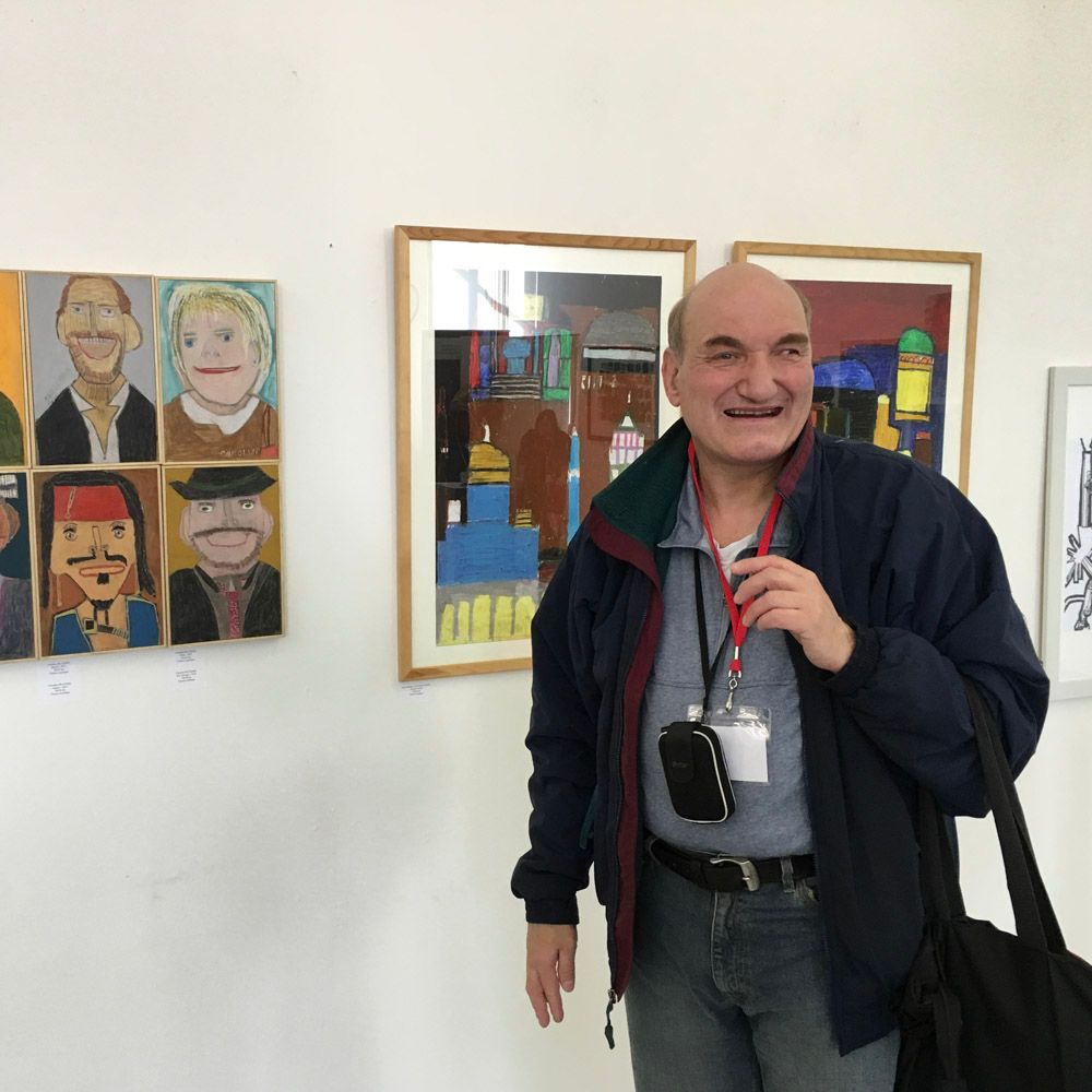 Man smiling in an art gallery, next to his colorful paintings. He's wearing a blue jacket and holding a bag.