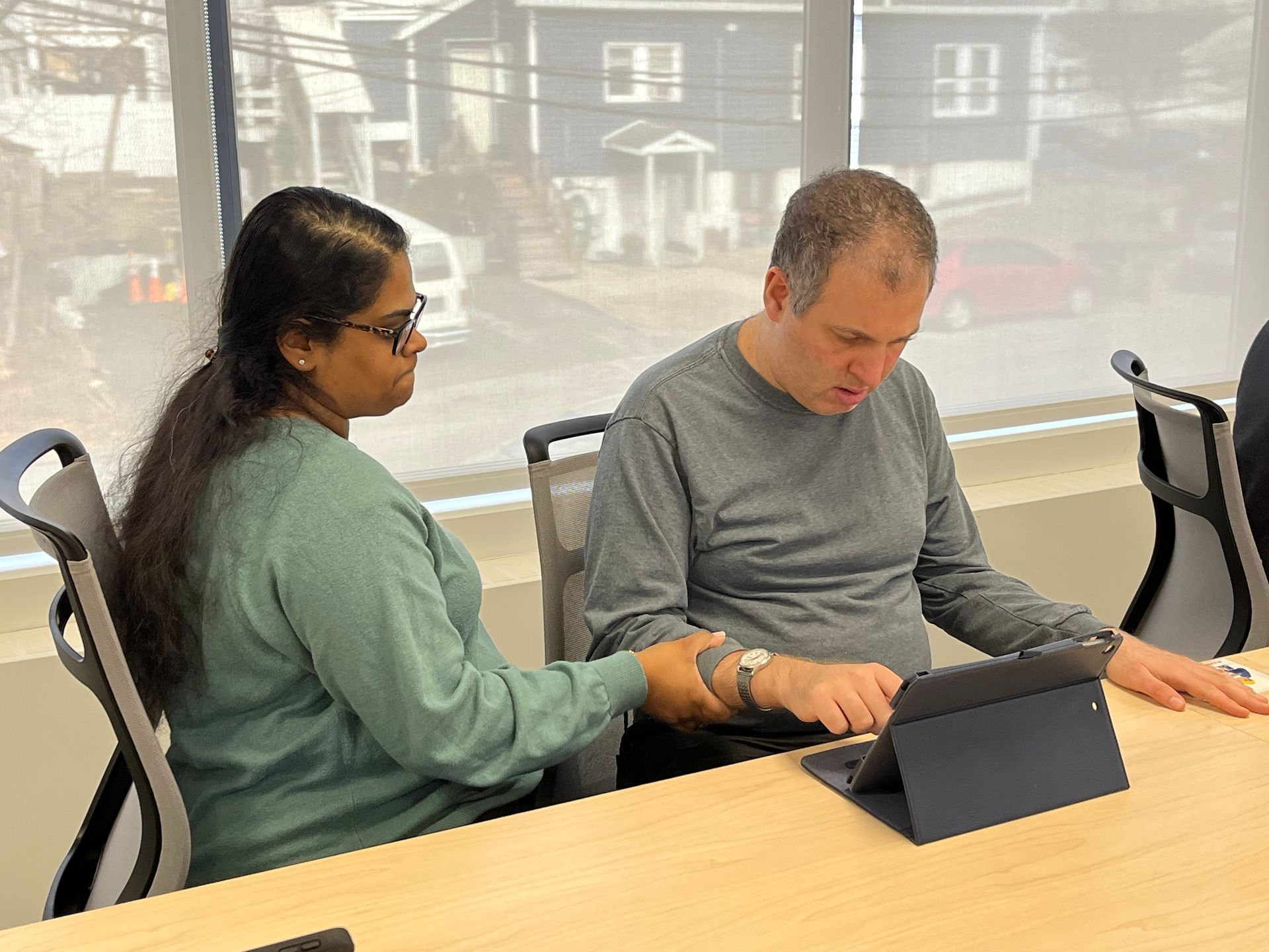 Woman assisting a man using a tablet at a table. Both are seated, inside a room with a window.