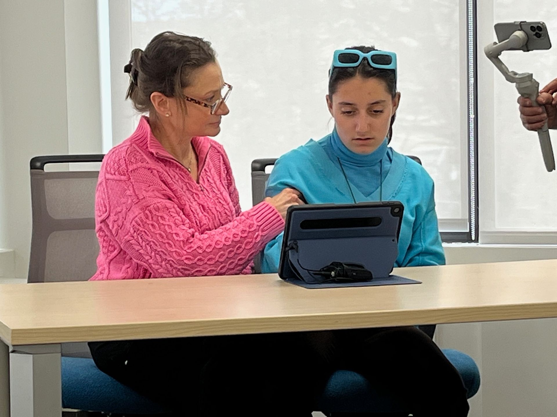 Woman in pink sweater assists a girl in blue looking at a tablet, held on a table. A phone on a gimbal records them.