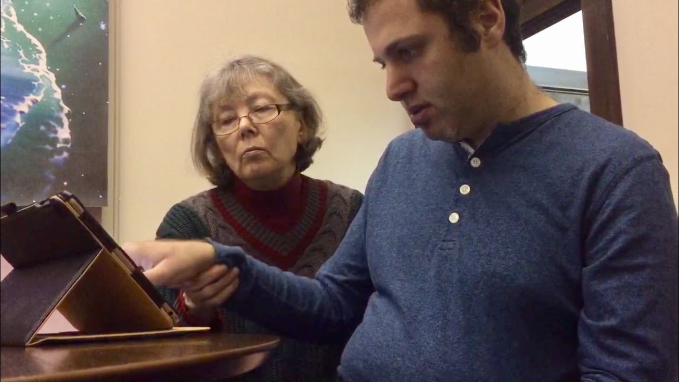 Man points to tablet screen, showing it to woman. Both are indoors, looking at the tablet.