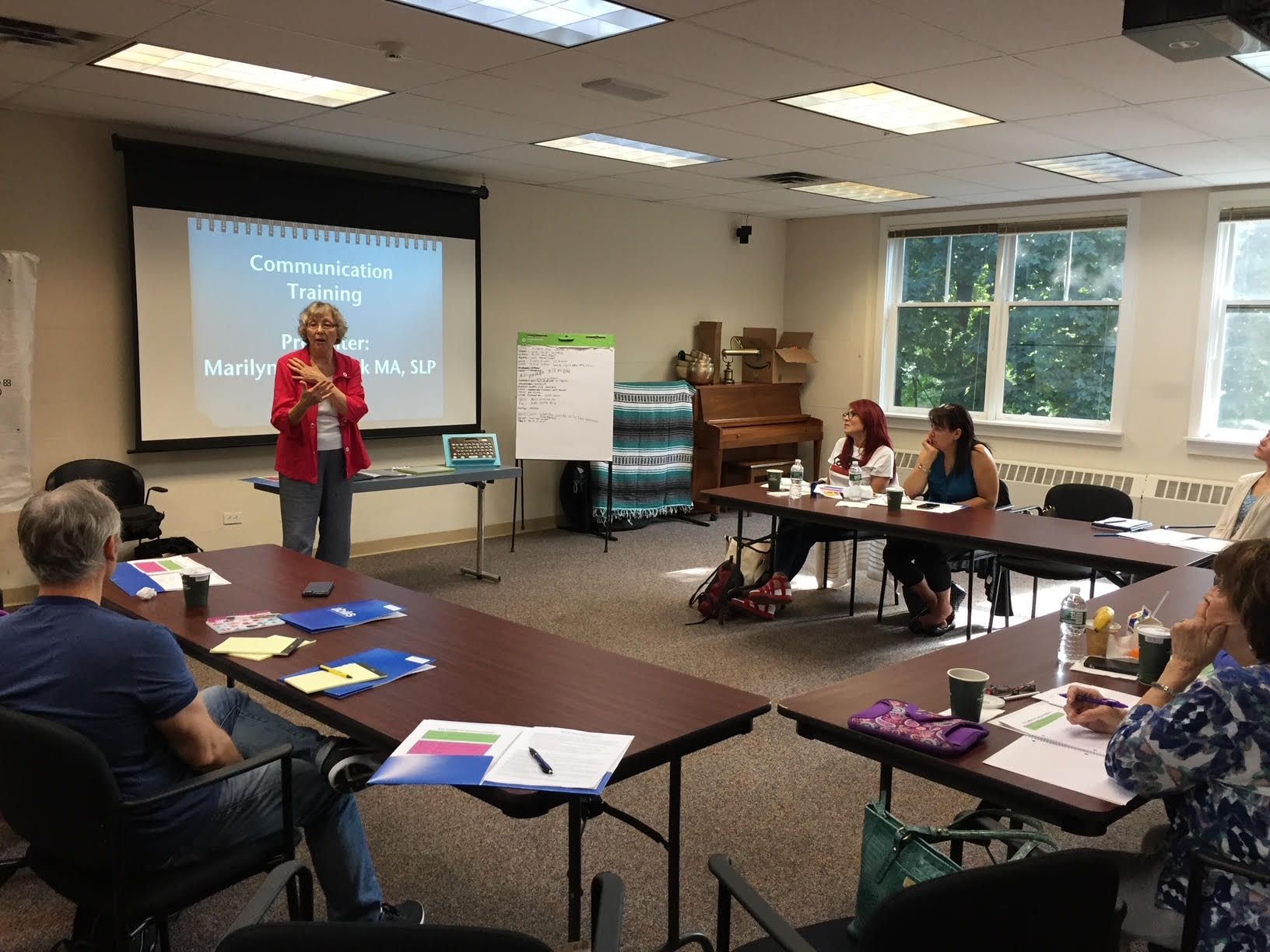 A woman presents in a classroom. Attendees sit at tables; a projector screen displays text.