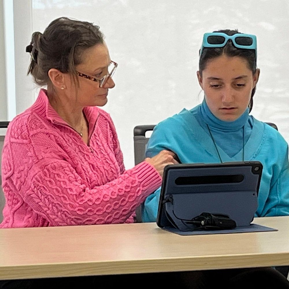 Woman in pink sweater assists another with blue glasses and tablet at a table.