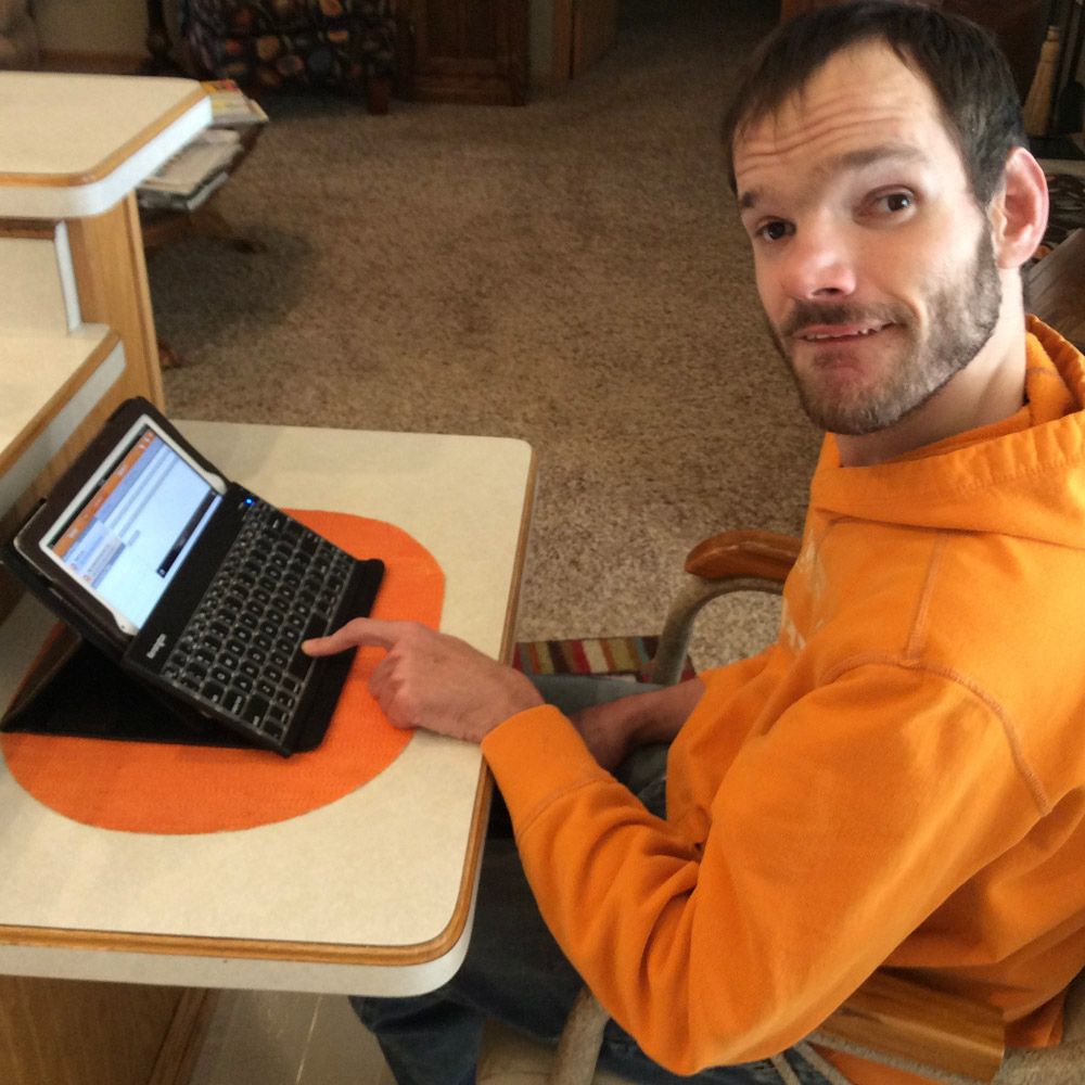 Man in orange hoodie seated, pointing at a laptop on a small table, interior setting.