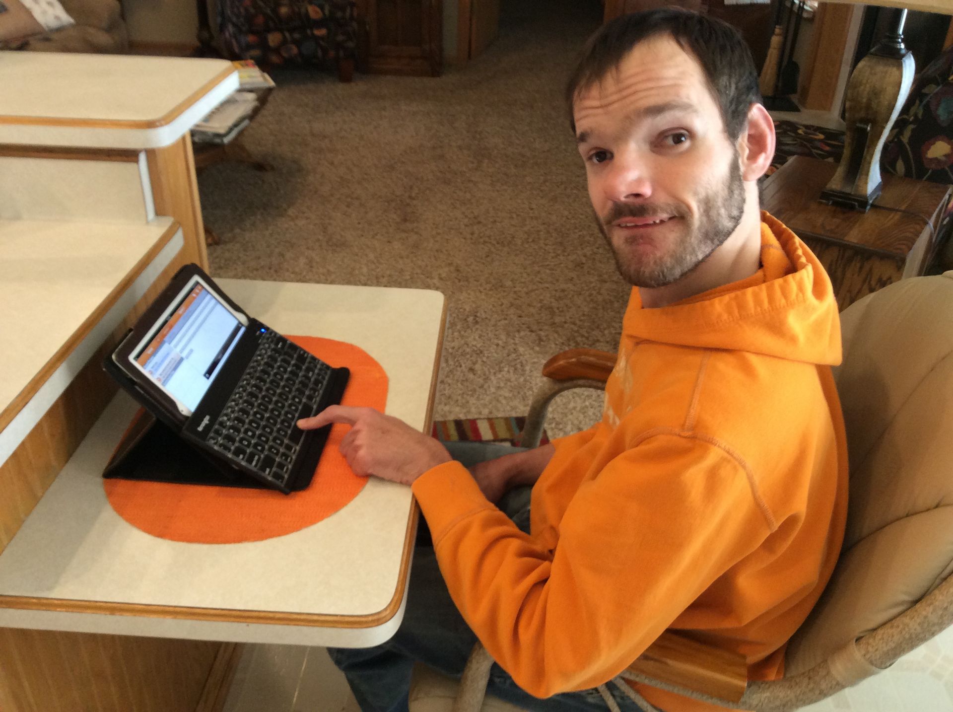 Man in orange hoodie seated at a desk, pointing to a laptop. Brown carpet in the background.