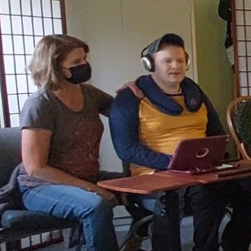 Woman in mask, touches man’s shoulder. Man wears headset, uses laptop. Sitting indoors.