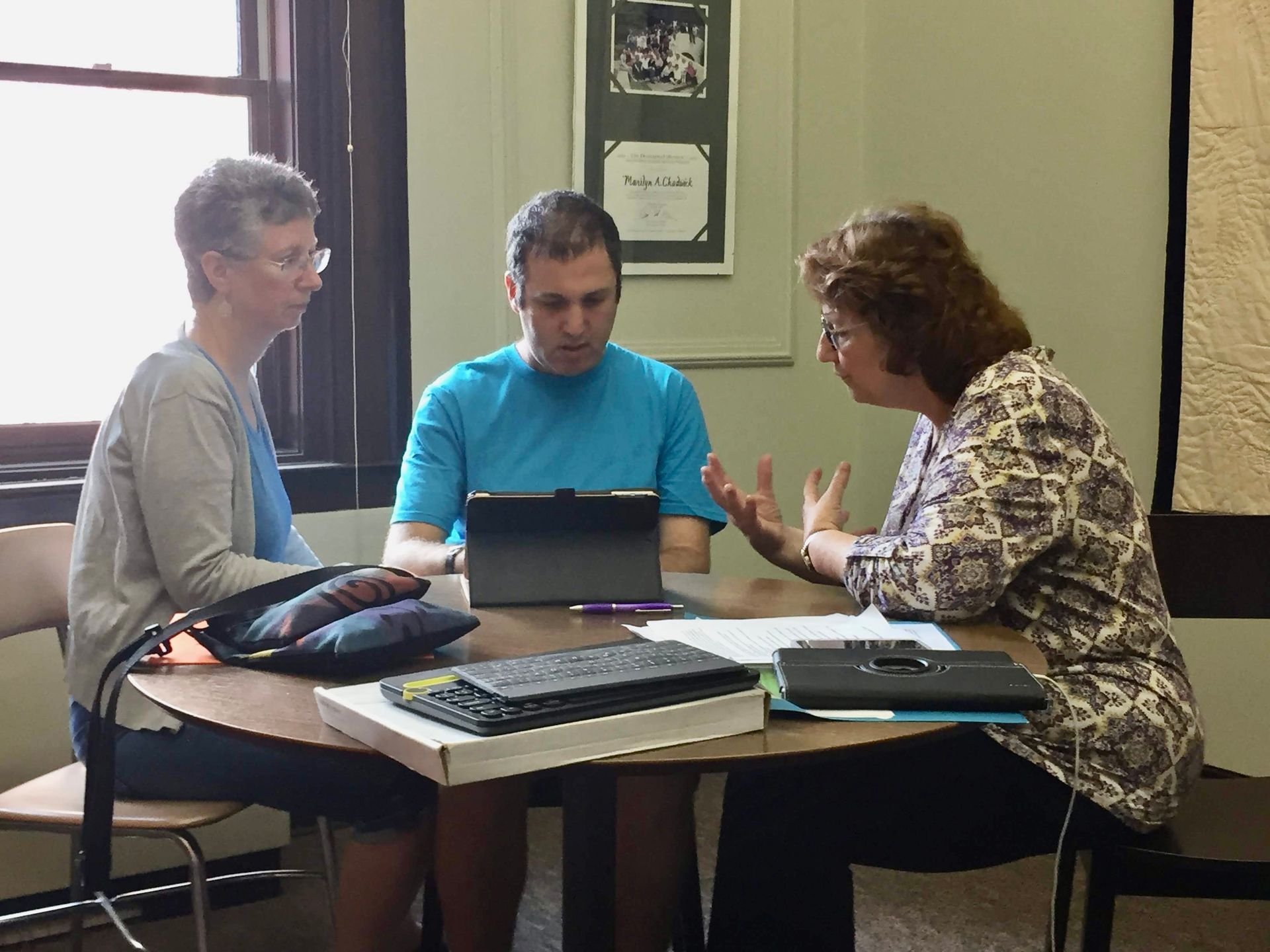Three people at a table, discussing. Man in blue shirt uses laptop. Woman gestures, woman on left observes.