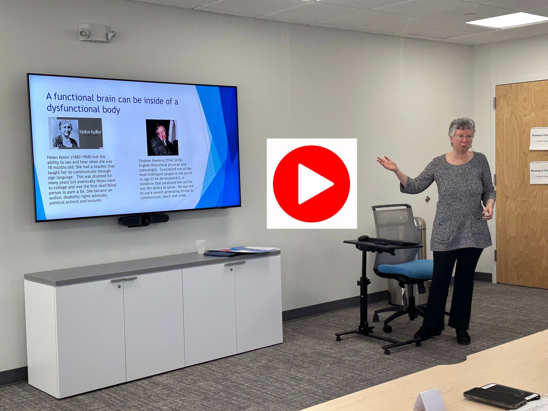 Woman presents in a conference room; a screen shows a presentation. A red play button overlays the scene.