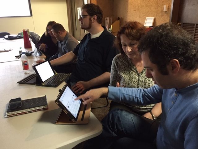 People in a meeting, some pointing at tablets on a round table. Indoor setting, natural light.