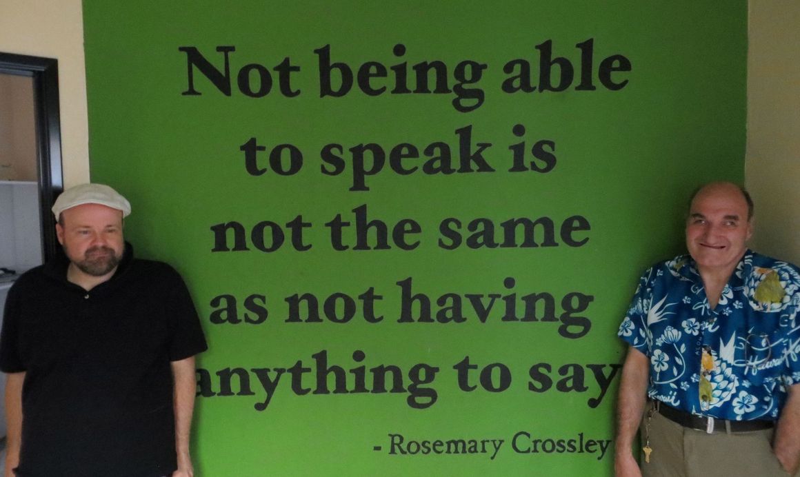 Two men stand next to a green wall with the quote: