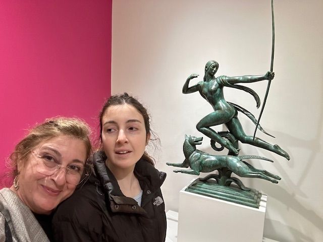 Two women pose by a bronze sculpture of a figure with a bow and dog in an art gallery.